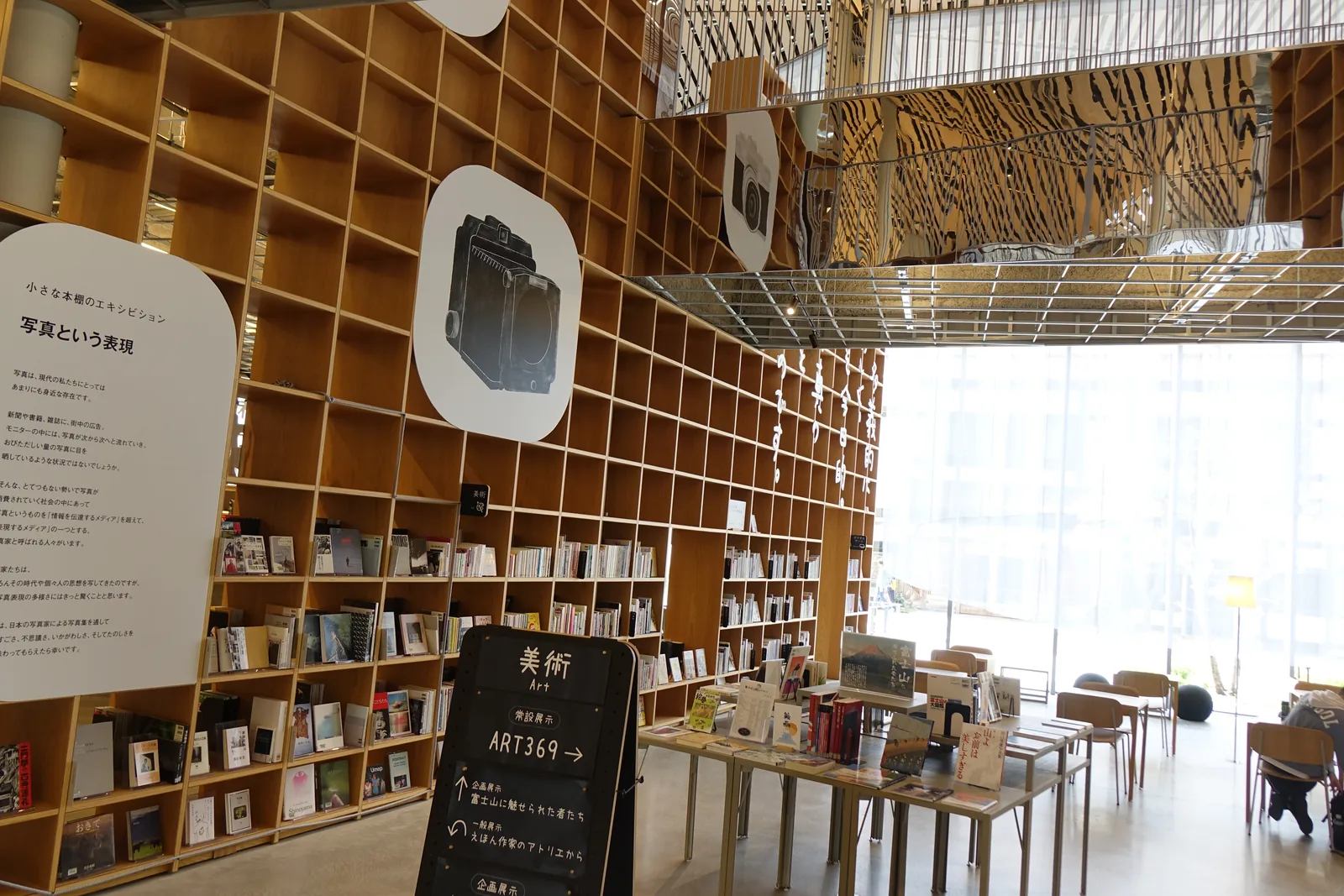 一樓藝術類書架前的展示區，木格書架上陳列著攝影與藝術書籍，書架上掛著圓形的標示與說明板，前方立著寫有美術與 ART369 的黑色指示牌，右側是玻璃立面旁的桌椅