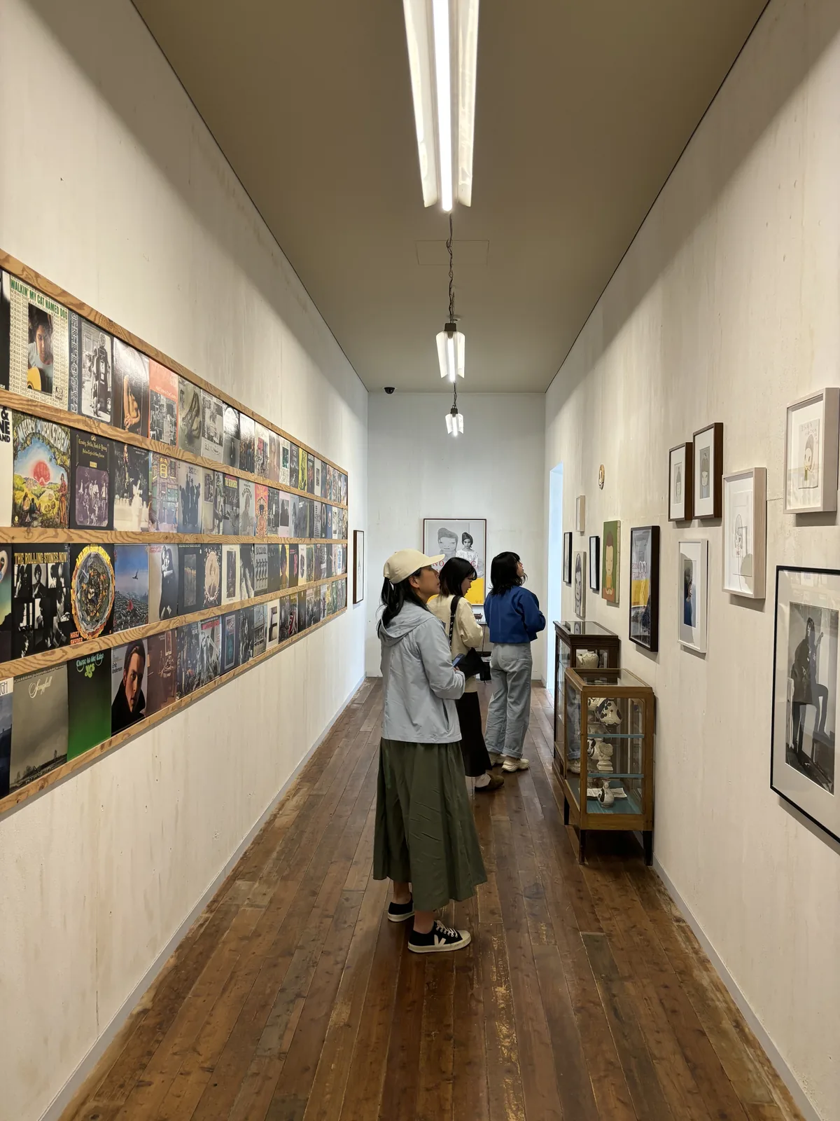 一條狹長的展示走廊，左牆上以木條架層層排列著唱片封套，右牆掛著裱框的照片與畫作，地板是深色的舊木地板，天花懸吊著一支日光燈，走廊盡頭的牆上掛著一幅畫