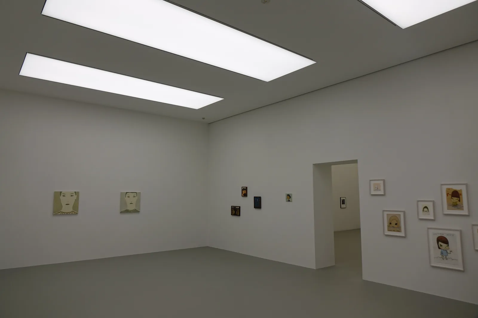 展示室一，白色的牆面與淺灰色的地坪，天花上嵌著兩條長方形的發光面板，牆上掛著數幅小型畫作，右側牆上開著一個通往下一室的開口
