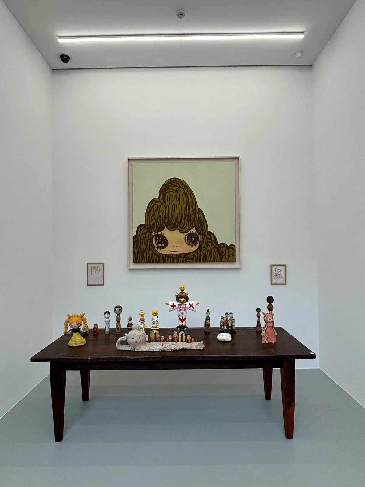 展示室三的內部，白牆上掛著一幅大型畫作，畫中是一個頭髮蓬鬆的女孩，畫下方是一張深色的木桌，桌上排列著小型人偶與物件，地坪是淺灰色