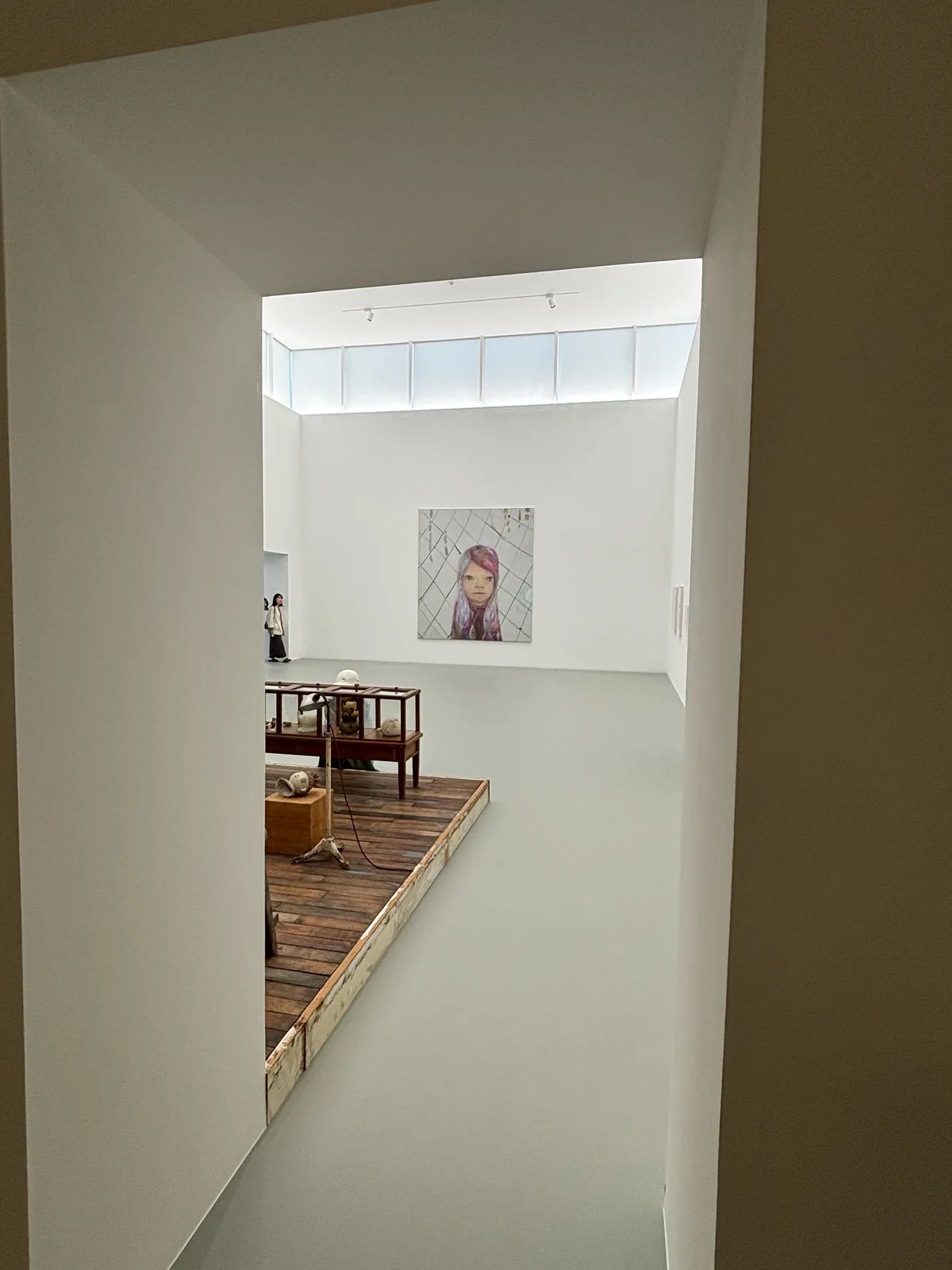 自一個深色牆面的開口望進展示室四，開口後方是挑高的白色空間，牆上方有一圈帶狀的高側窗，遠處牆上掛著一幅畫，左下方可見一塊舊木板平台與木桌