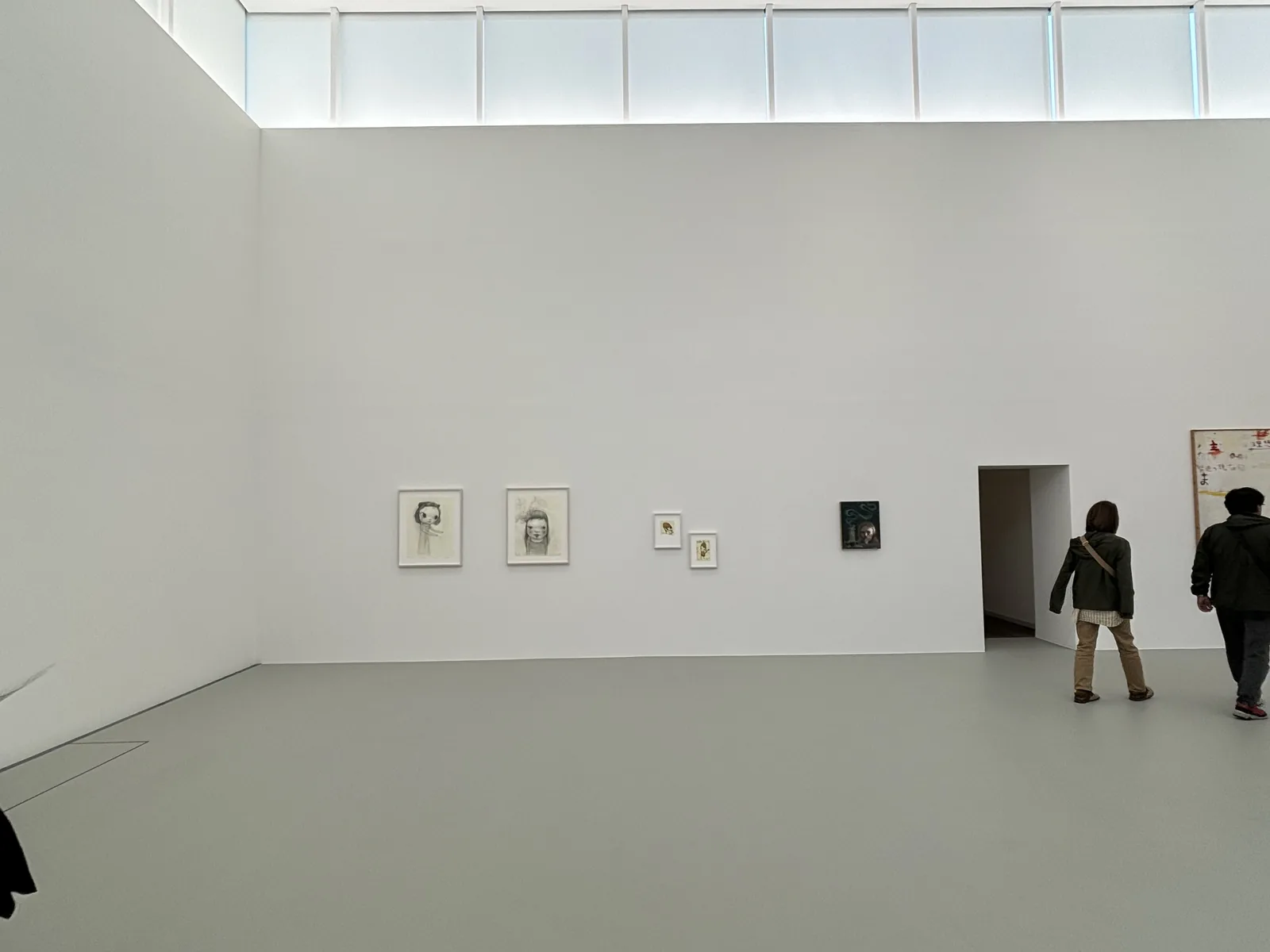 展示室四，四面白牆的上方各有一圈帶狀的高側窗，窗外的光均勻地照亮牆面，牆上掛著幾幅小型的素描與畫作，右側的開口通往下一室，有兩位參觀者正走進去
