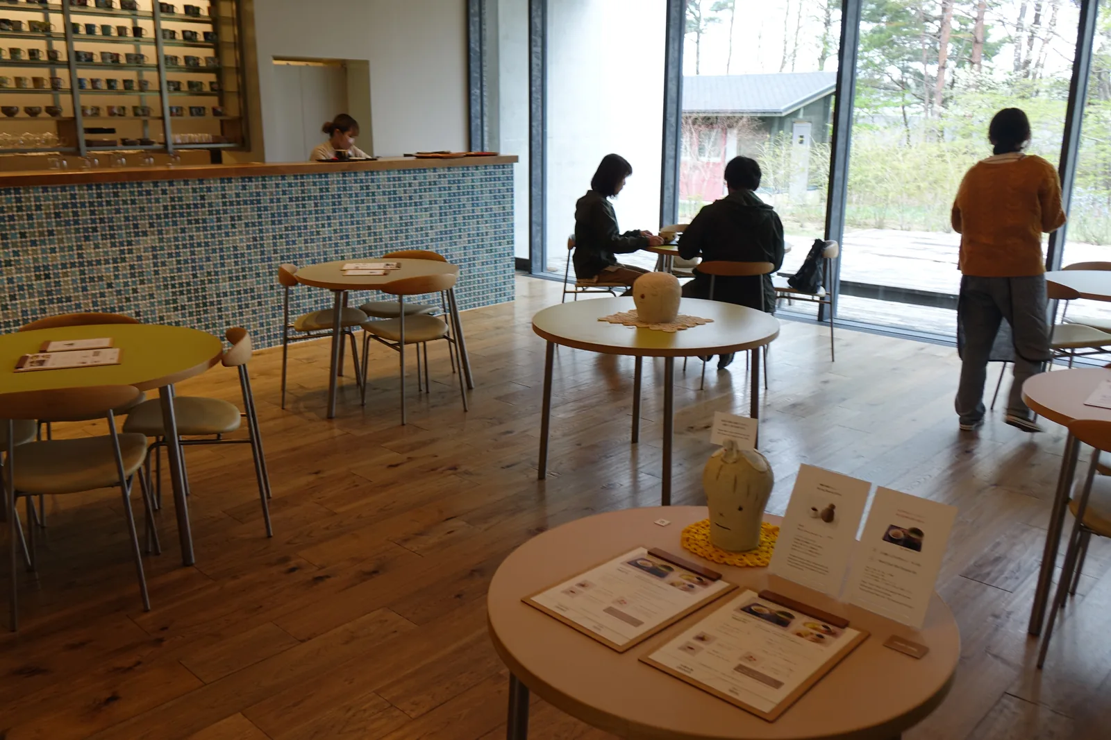 Konara Cafe 的內部，木地板上擺著幾張圓桌與金屬腳的椅子，左側是貼著藍色小磁磚的吧台，吧台後方的牆上嵌著一排木格，右側是落地玻璃窗，窗外是樹林與一棟小屋