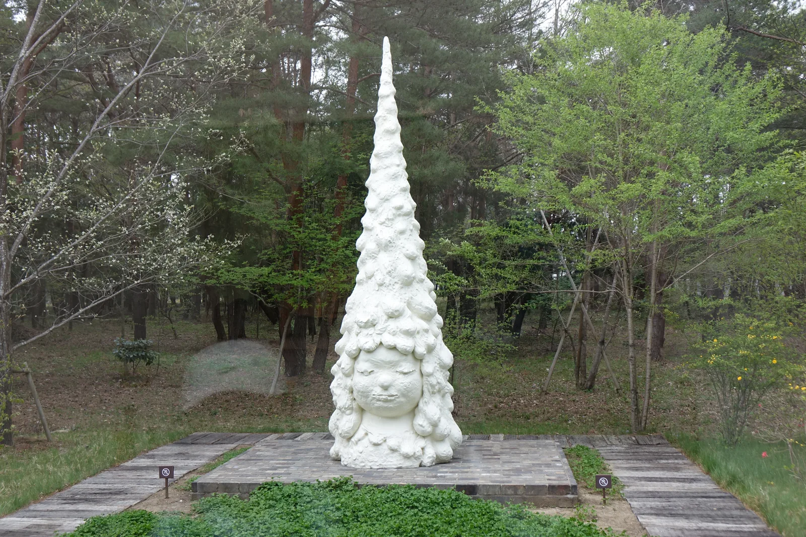 庭園中的白色雕塑，一座高聳的圓錐形雕塑立在方形的木板台座上，雕塑表面布滿重疊的頭像，底部是一張女孩的臉，四周是草坪、木板步道與樹林