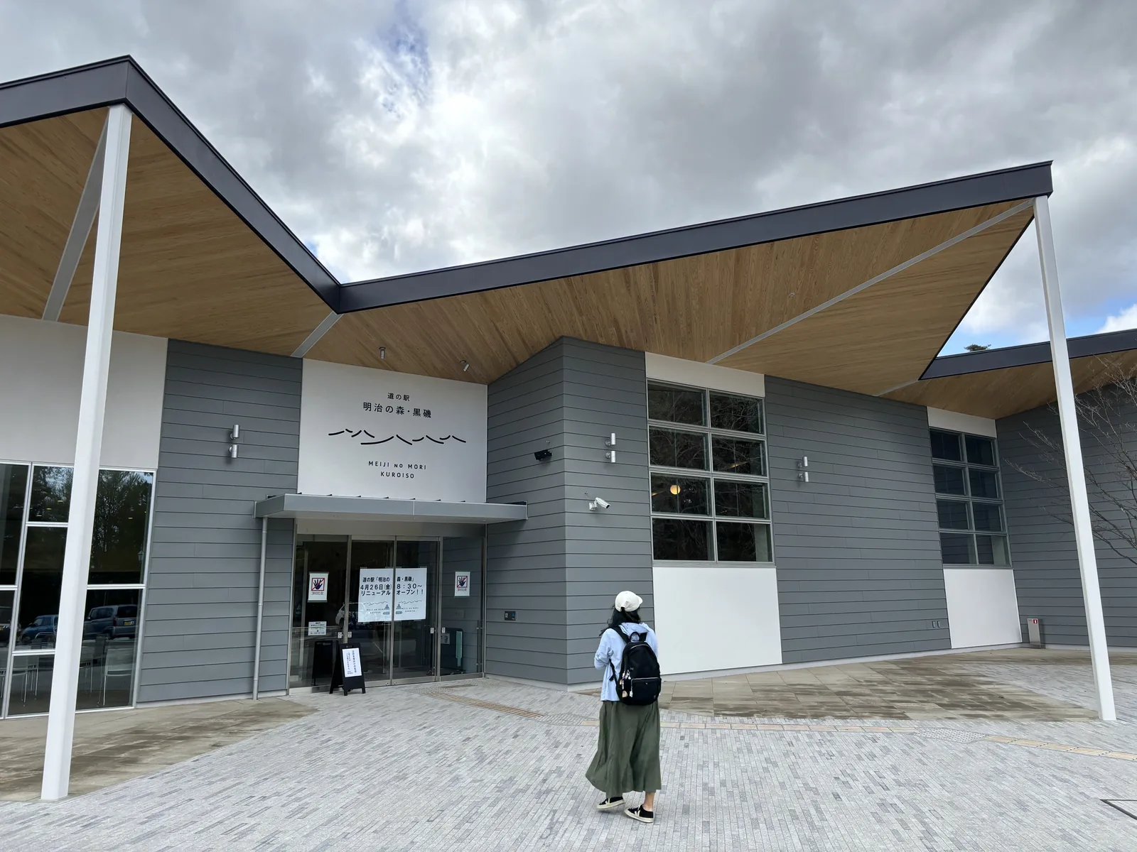 公路休息站明治之森・黑磯的新館，灰色橫向壁板的立面上方是一片向外伸出、底面為木板的折板狀大屋簷，以白色細柱支撐，入口上方掛著館名，前方是鋪石的廣場