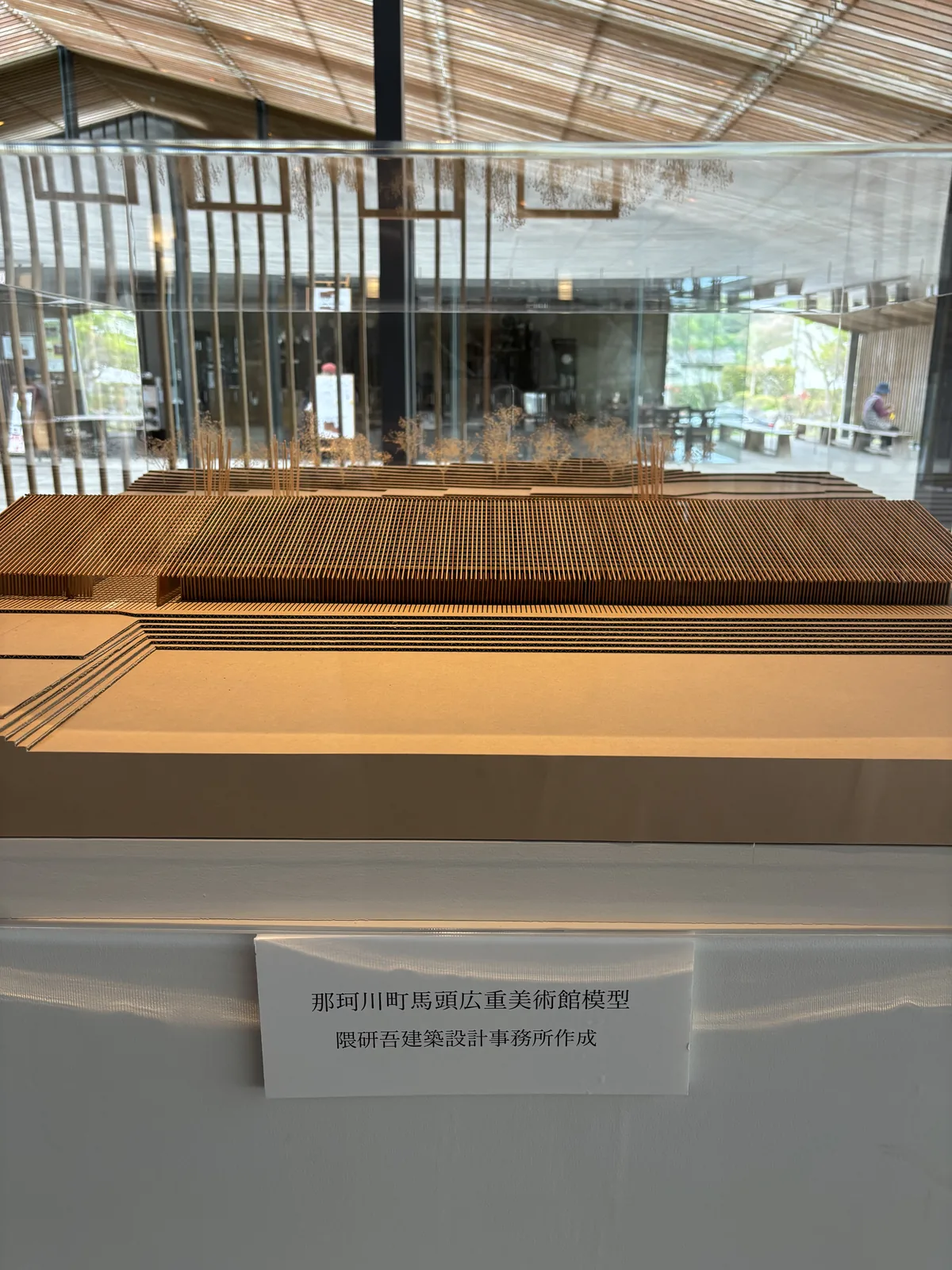 建築模型，以木條排列出覆蓋整棟建築的切妻屋頂格柵，模型的一側是幾層階梯狀的台地，台座上貼著寫有館名與設計事務所名稱的標籤，背景是透過玻璃可見的館內與中央通道