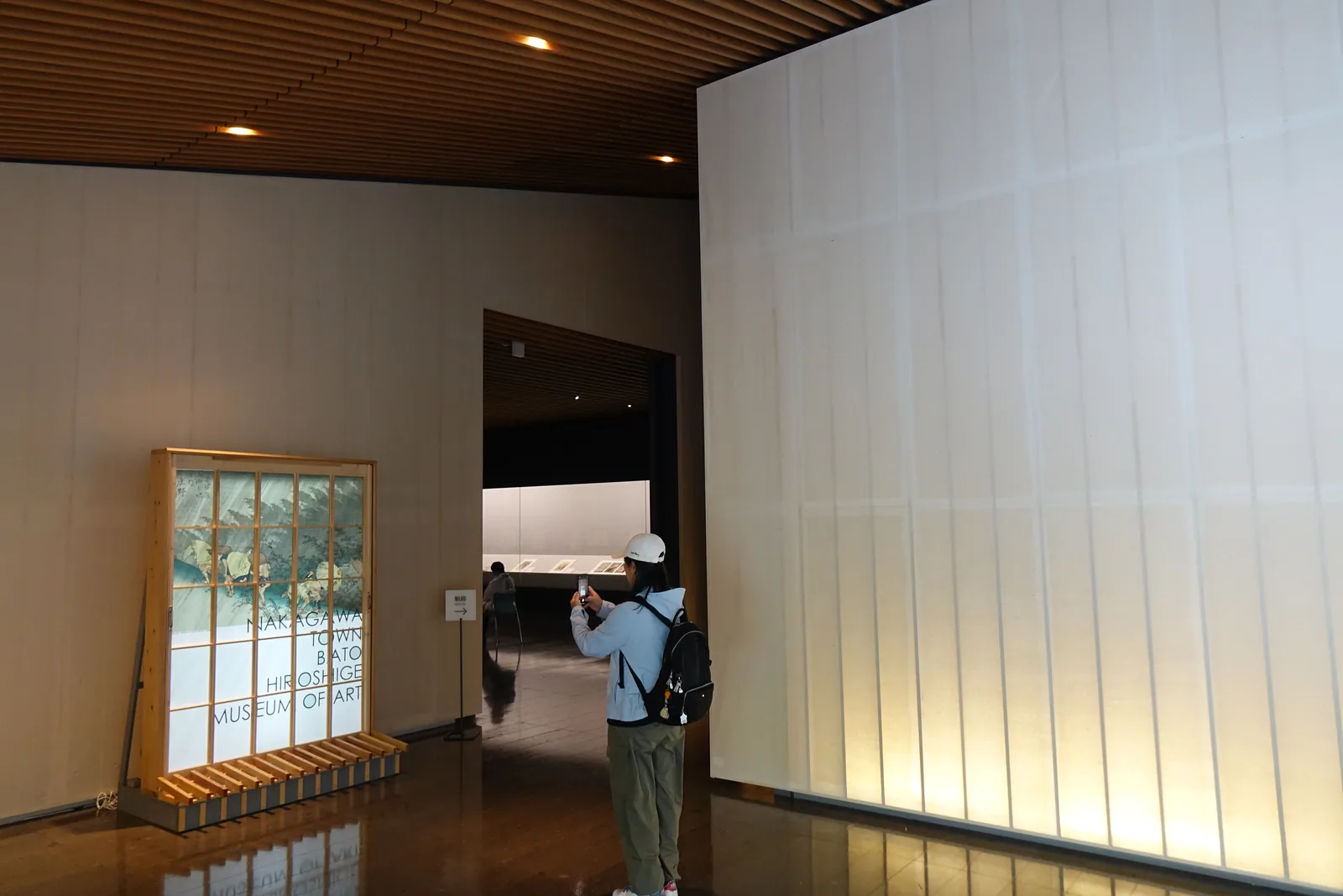 展示室入口前的前室，左側牆前立著一面木格框架的館名看板，中央是通往展示室的暗色開口，開口內可見展示櫃的光，右側是一整面發亮的和紙牆，一位參觀者正在拍照