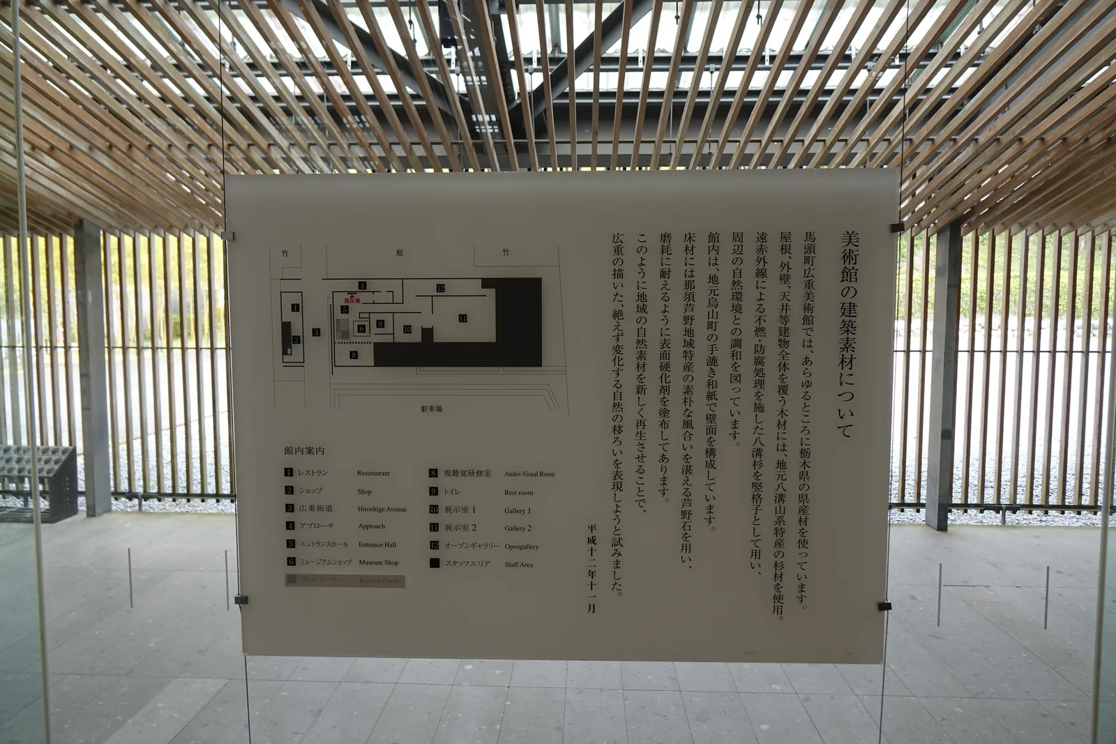 館內的說明板，以直書的文字說明美術館的建築材料，左側是一張館內平面圖與設施清單，說明板以細索懸吊在玻璃前，背景是格柵天花與杉木直格柵
