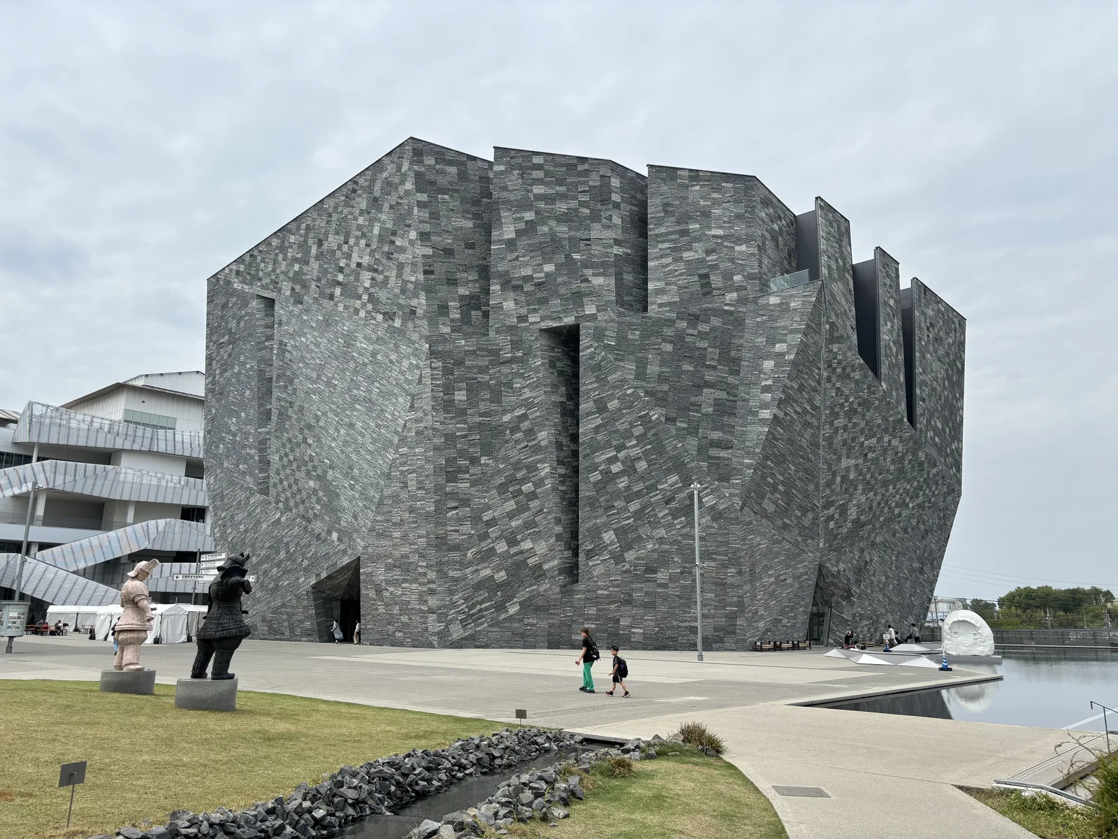 角川武藏野博物館外觀，花崗岩包覆的多面體量體立在草坪的盡頭，頂部被切成數道鋸齒狀的缺口，前方是以黑色碎石鋪成的細長水路與步道