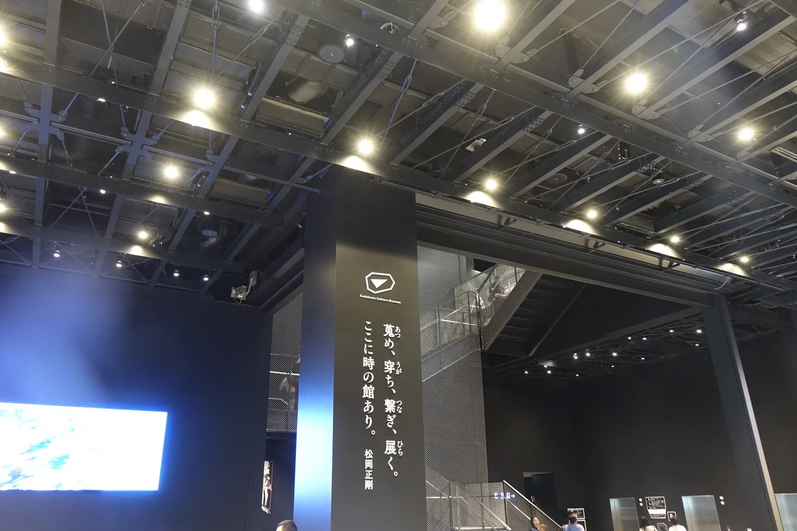 二樓入口大廳，天花是外露的黑色鋼構與成列的點狀照明，正面是一面細長的螢幕，左側是黑色的牆面與玻璃，地面是淺灰色的石材，大廳中有排隊的人與欄柱