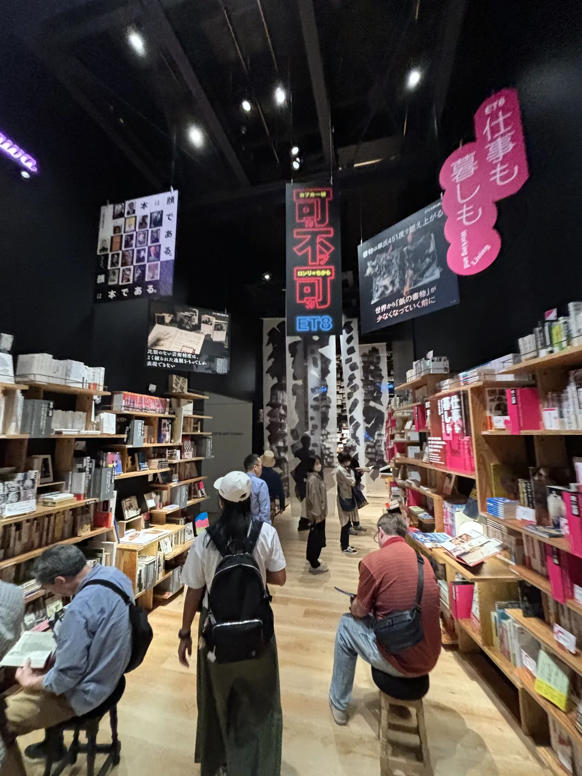 Book Street 的另一段，左右兩側是木製書架與紅色的書架，上方懸吊著寫有大字的紅色看板與黑色的布條，有人坐在書架旁閱讀