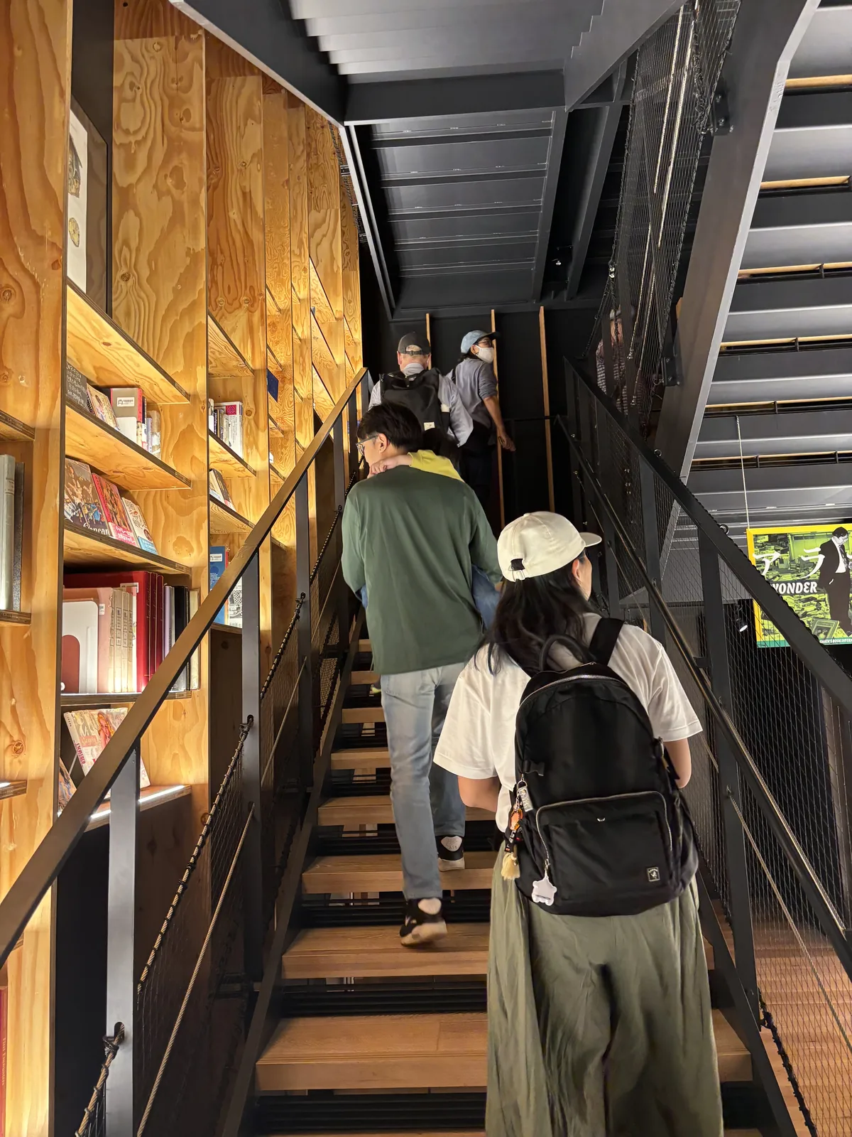 通往五樓的樓梯，左側是木製的書架與合板的牆面，書架中排列著書，右側是黑色的鋼製樓梯與欄杆，幾位參觀者正在上樓