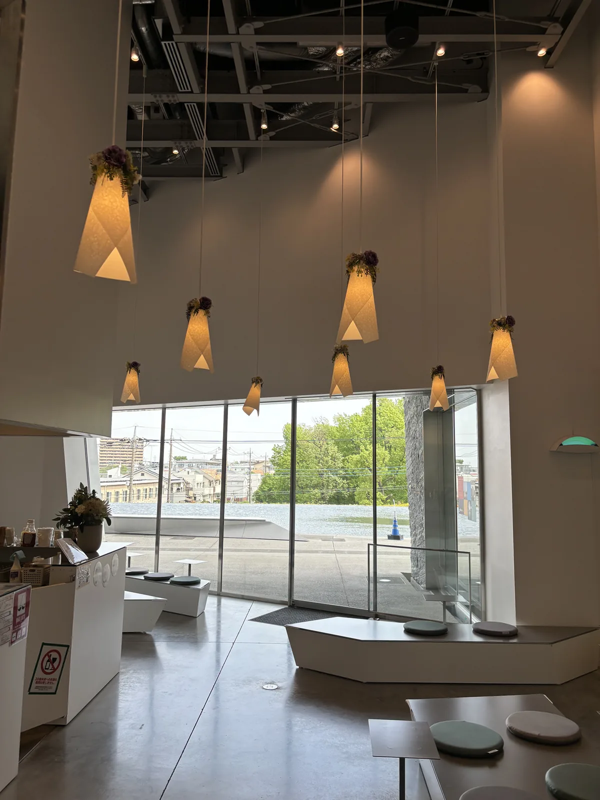 二樓 KadoCafe 的店內，天花懸吊著數盞白色的紙燈罩吊燈，燈罩上方以乾燥花裝飾，正面是整面的落地玻璃，窗外是平台與樹木，室內是低矮的白色座台與圓形坐墊