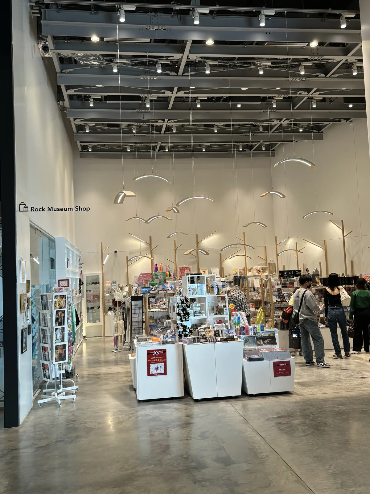 二樓 Rock Museum Shop 的店內，白色牆面的高大空間中立著數座白色的陳列台，天花是外露的鋼構，懸吊著白色的弧形燈具，店內有許多購物的人