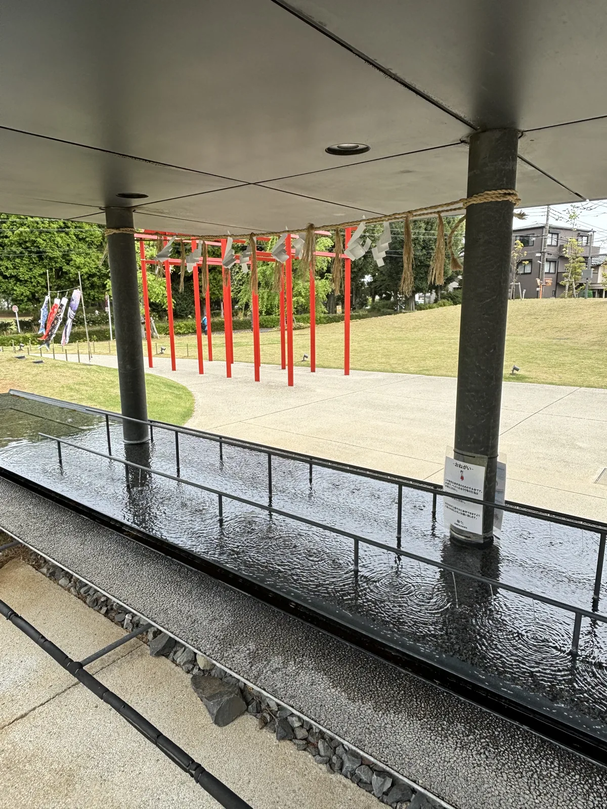 自神社的屋簷下望向側院，前景是一條以黑色石材鋪成的水路，屋簷下立著兩根黑色的柱子，柱間掛著注連繩，側院的草坪上排列著紅色的鳥居