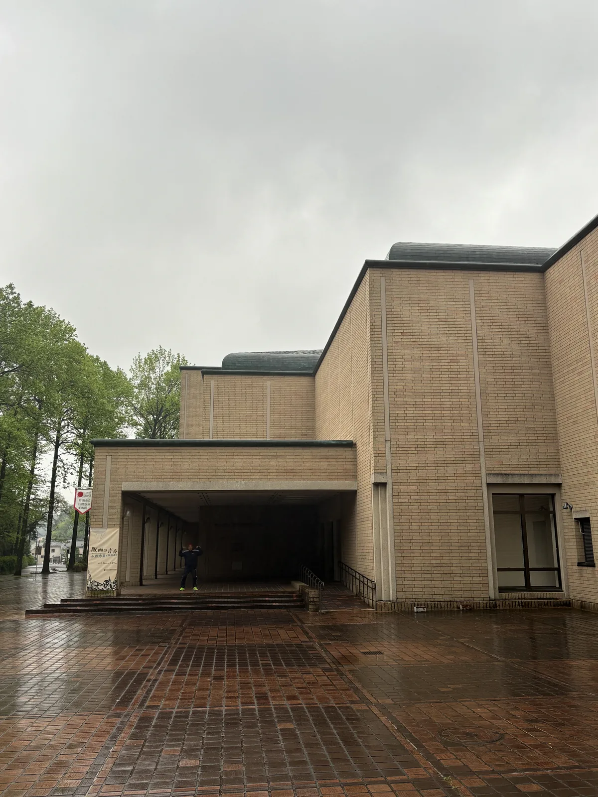 町田市立國際版畫美術館的入口一側，米色磚牆的量體層層後退，左側是低矮寬深的雨庇，雨庇下有一人站立，前方是被雨淋濕的磚鋪廣場，屋頂上方露出綠色的金屬屋面