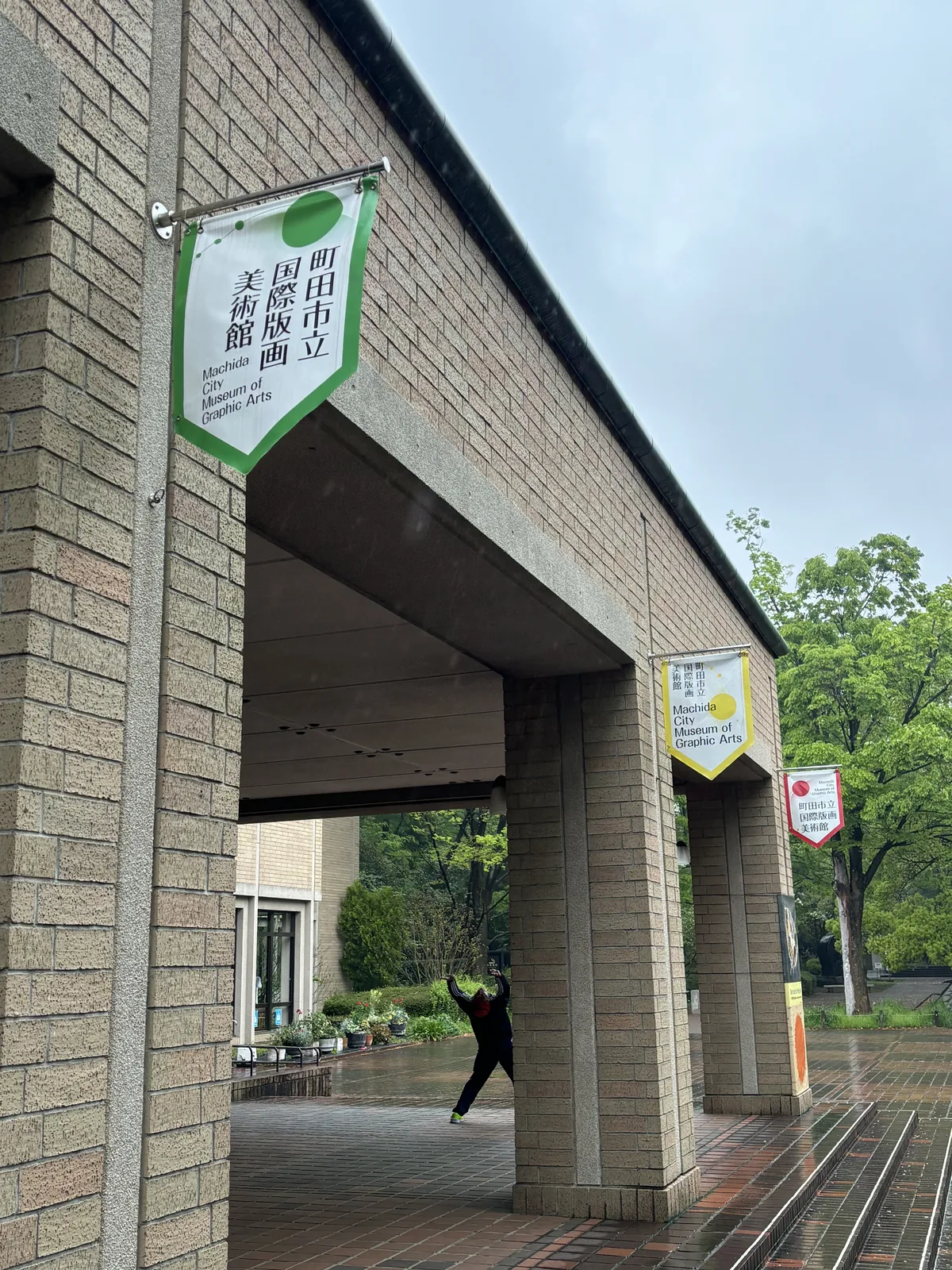 列柱廊的近景，磚包覆的方柱承接灰色的大樑，柱上懸掛綠色與黃色的館名旗，廊下陰影中可見遠處的庭木與植栽