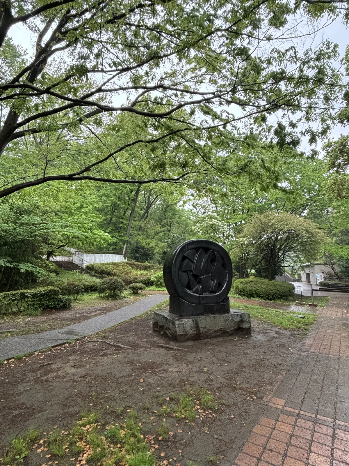 公園內的步道旁，一件黑色的圓盤形雕塑立在石座上，周圍是草地、灌木與高大的落葉樹，步道以紅磚與石材鋪設