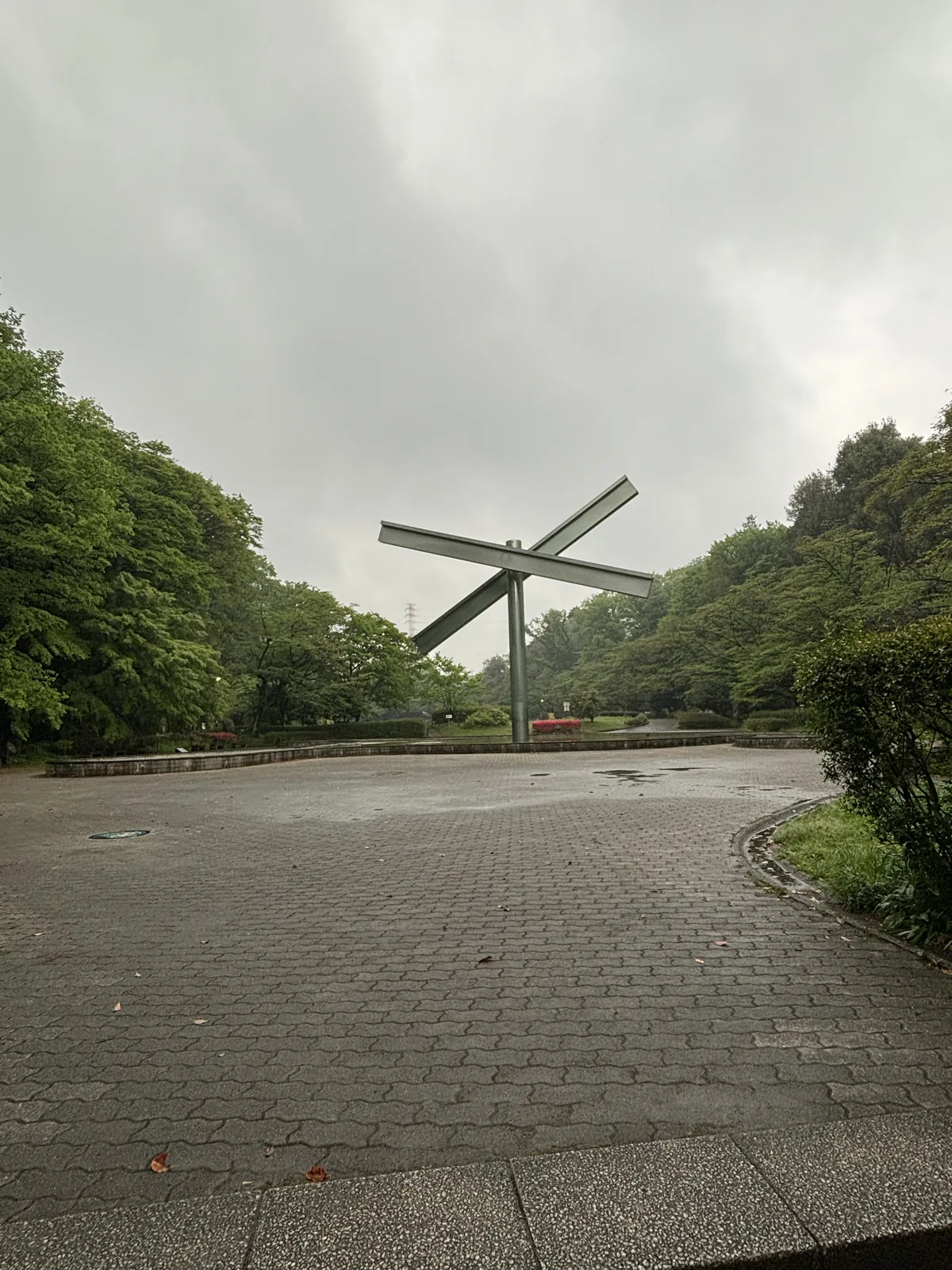 雨中的公園廣場，地面鋪著方形的石磚，中央立著一件由兩根鋼構斜交成 X 形的大型雕塑，周圍是濃綠的樹林