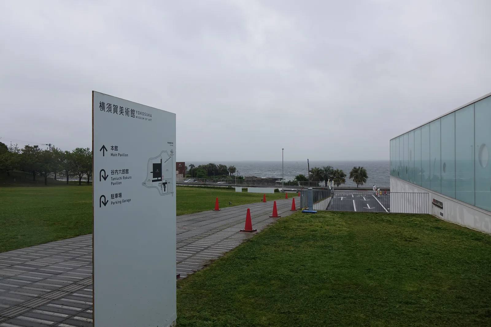 自海側的道路走向美術館，左側是一面館名的指示牌與草坪，右側是一棟玻璃包覆的低矮量體，量體前方是一條鋪石的步道，遠處是陰天下的海