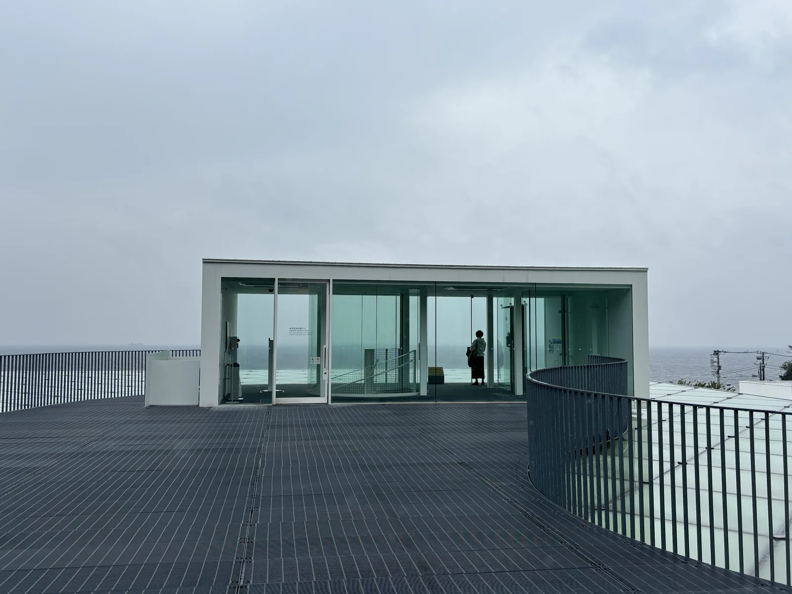 屋頂廣場，深灰色的 grating 鋪面上立著一座白框的玻璃小屋，小屋內有人影，右側是一道弧形的金屬欄杆，背景是陰天下的海