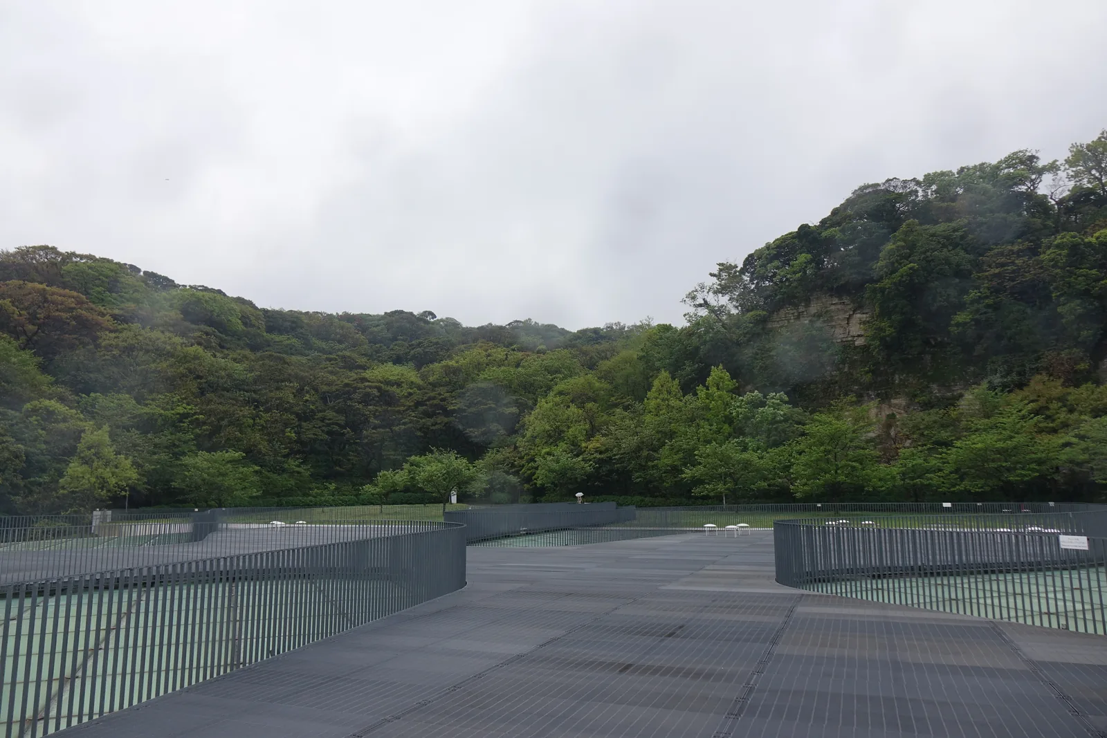 自屋頂廣場望向山側，深灰色的 grating 鋪面延伸至草坪，草坪的盡頭是被森林覆蓋的山坡，鋪面的邊緣是低矮的金屬欄杆