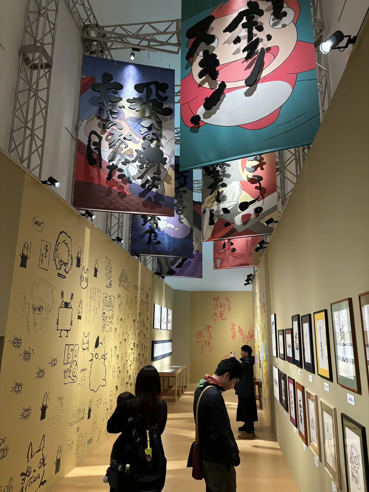 企劃展示室的高大空間，牆面以淡黃色的手繪圖案覆蓋，上方以鋼架懸吊著數面印有大字的布幕，下方有參觀者