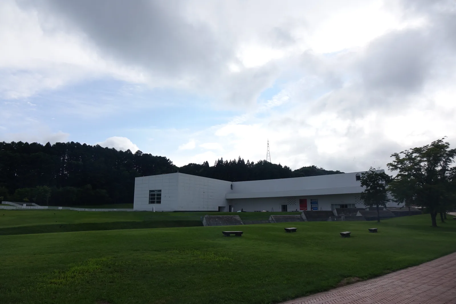 遠望美術館白色量體建築，草坪前景可見梯形淺色鋪面與草皮交錯的幾何切割，右下角紅磚步道