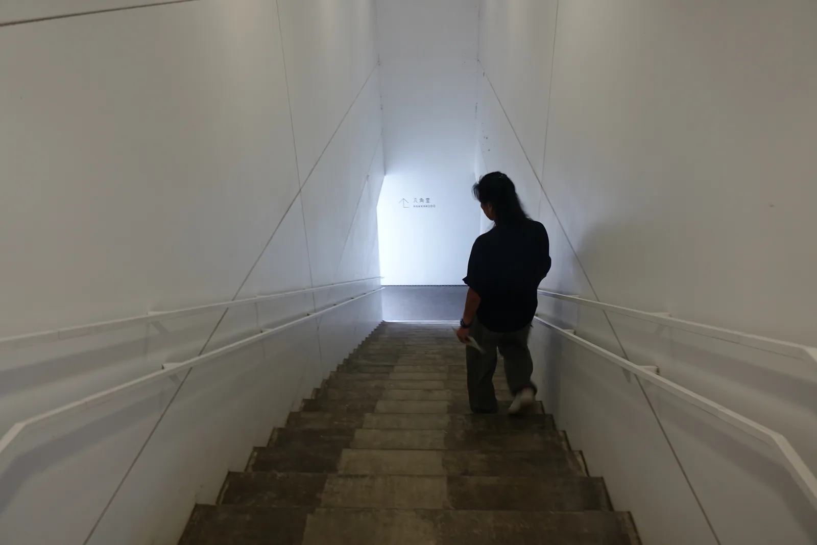 白色三角斷面斜天花隧道式階梯通道，兩側白牆向上收窄，遠端牆面標示八角堂指標