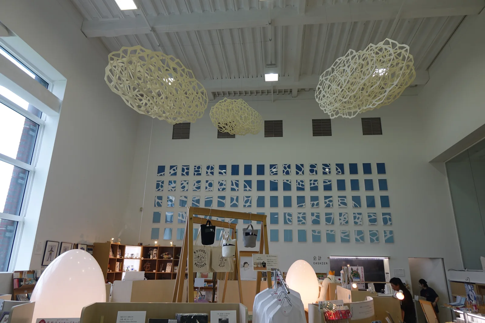 美術館商店室內全景，天花懸掛幾何鏤空紙質裝飾燈，牆面為藍白拼接方格圖樣，陳列托特包與書籍等商品