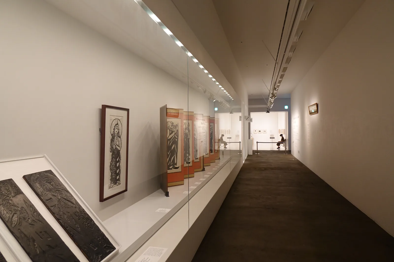 長廊型展示空間，左側玻璃展示櫃陳列棟方志功版畫與雕版原版，右側深色木質地板延伸至遠端展廳