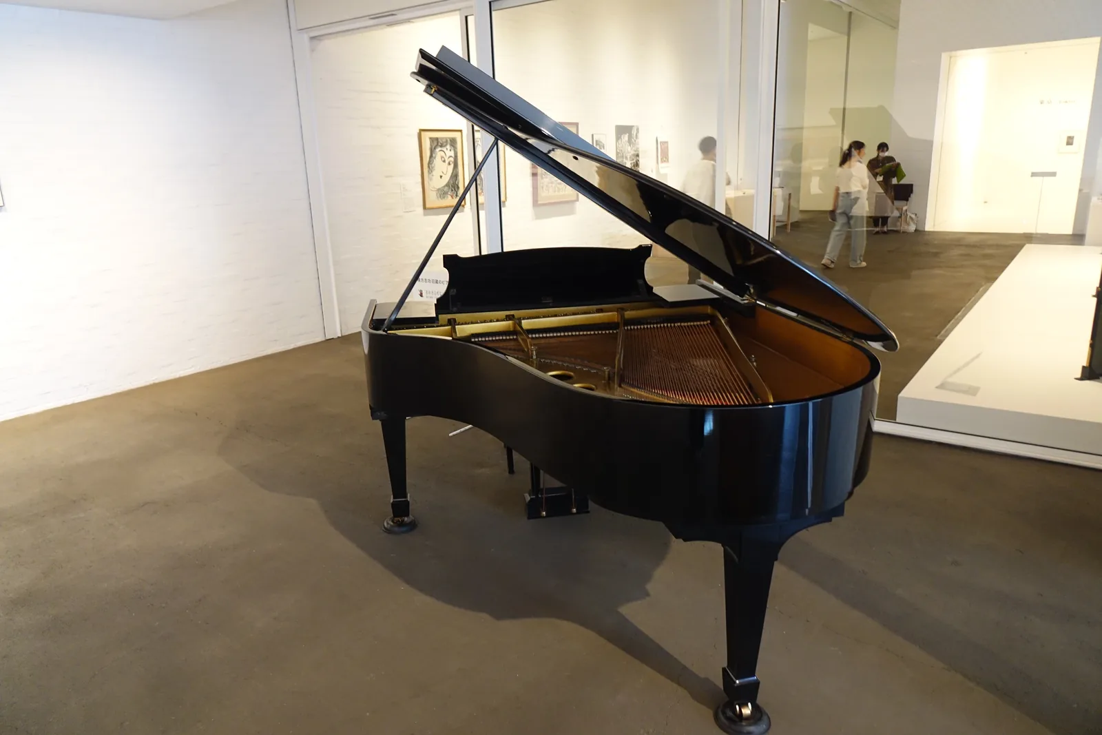 白色展示室內置放一架黑色平台鋼琴，深色地面，後方為展示牆與玻璃