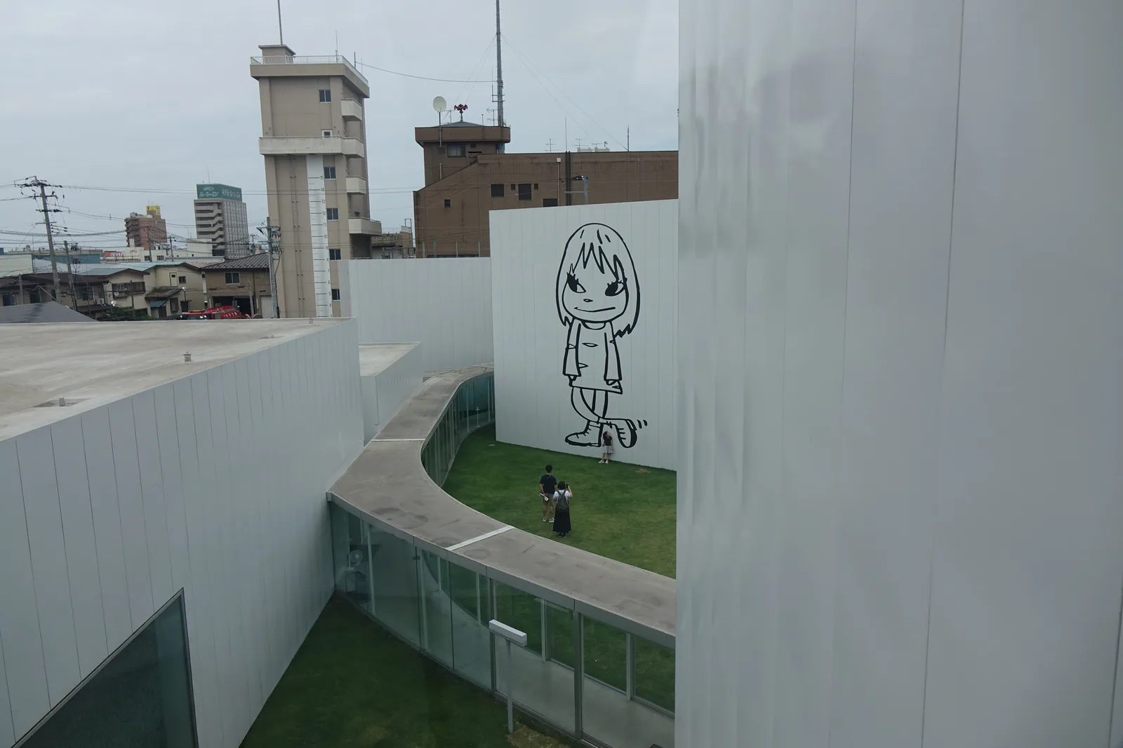 自樓梯間窗口俯視：弧形廊道的平屋頂繞過草坪，盡頭是巨大的線描女孩壁畫