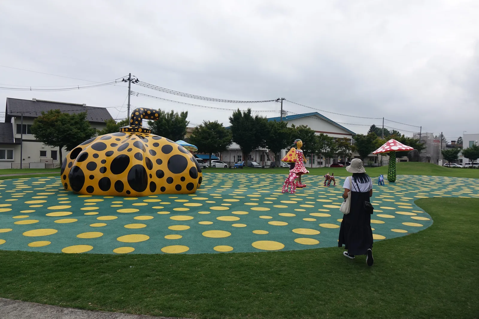 藝術廣場，綠色地面散布黃色圓點，其上立著黃底黑點的大型南瓜雕塑與其他作品，後方為街道與建築