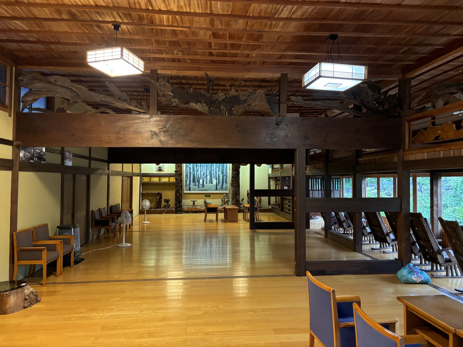 本館大廳：淺色木地板、木板天花與方形吊燈，橫梁上方嵌著原木雕刻，右側整排搖椅面向窗外