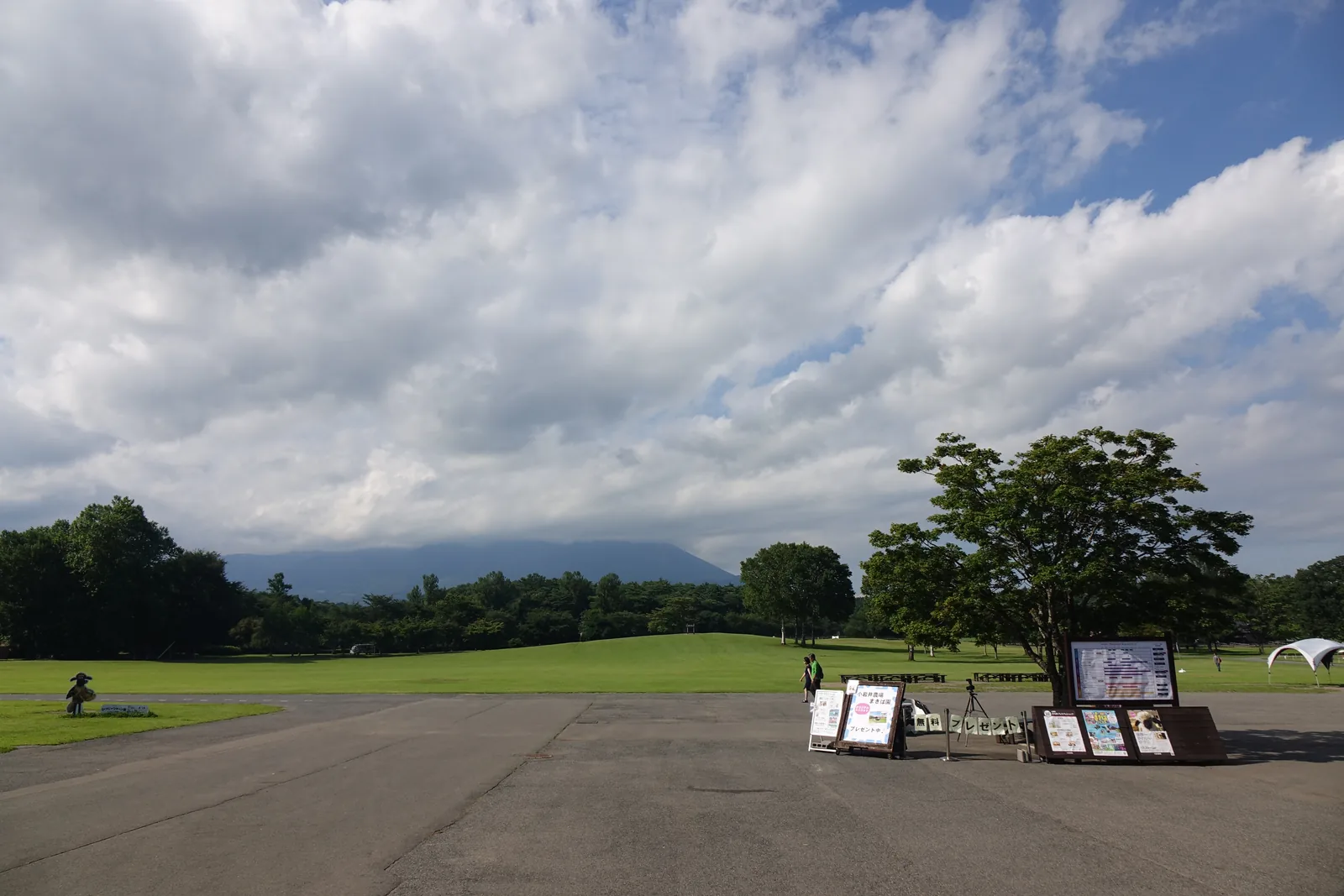 牧場園入口廣場：柏油廣場盡頭是草地與一株大樹，遠方樹林帶之上是被雲遮去一半的岩手山