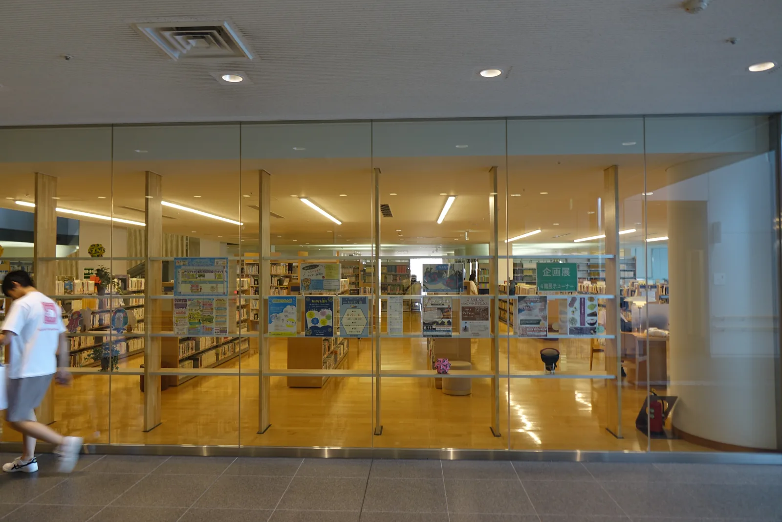 一間以玻璃牆面向走廊的室內，內部為木地板與書架，玻璃前有行人走過