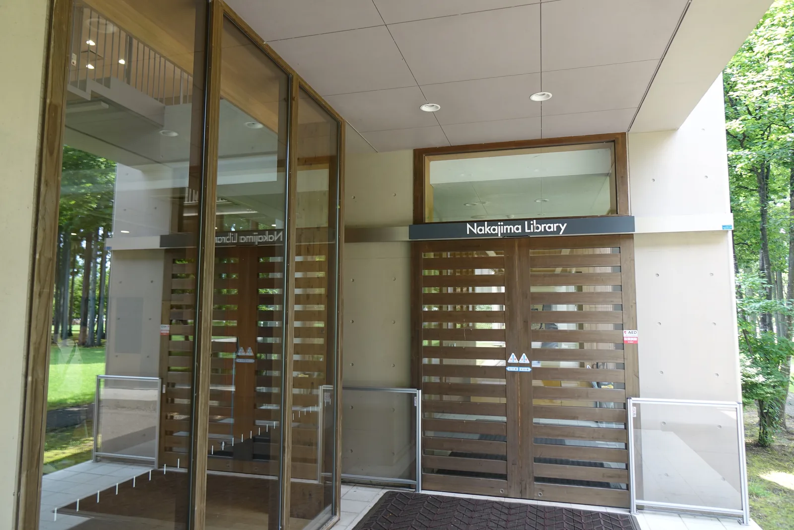 圖書館入口，淺色混凝土框架下為橫向木條構成的雙開門，門楣上掛深灰色標示板寫著Nakajima Library，左側為玻璃風除室