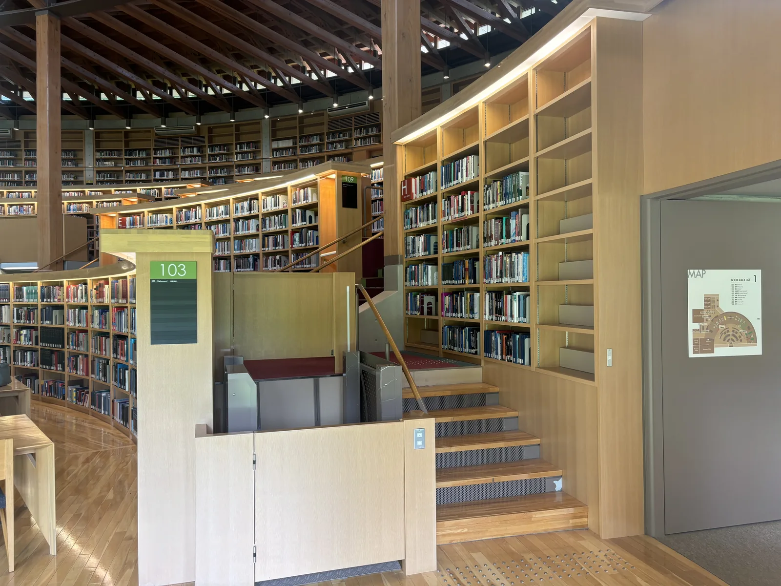 閱覽廳內一處樓梯，木踏板沿弧形書架拾級而上，書架背後為逐層升高的其他書架與上方的傘形屋頂，右側牆面貼著館內配置圖