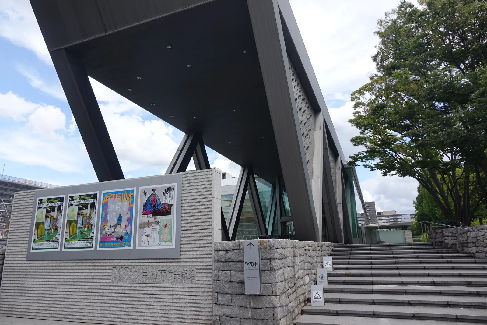 美術館臨街側入口，左側為張貼展覽海報的石材牆，右側為通往大廳的寬大石階，上方為深色挑簷與V字形斜柱，右側為行道樹