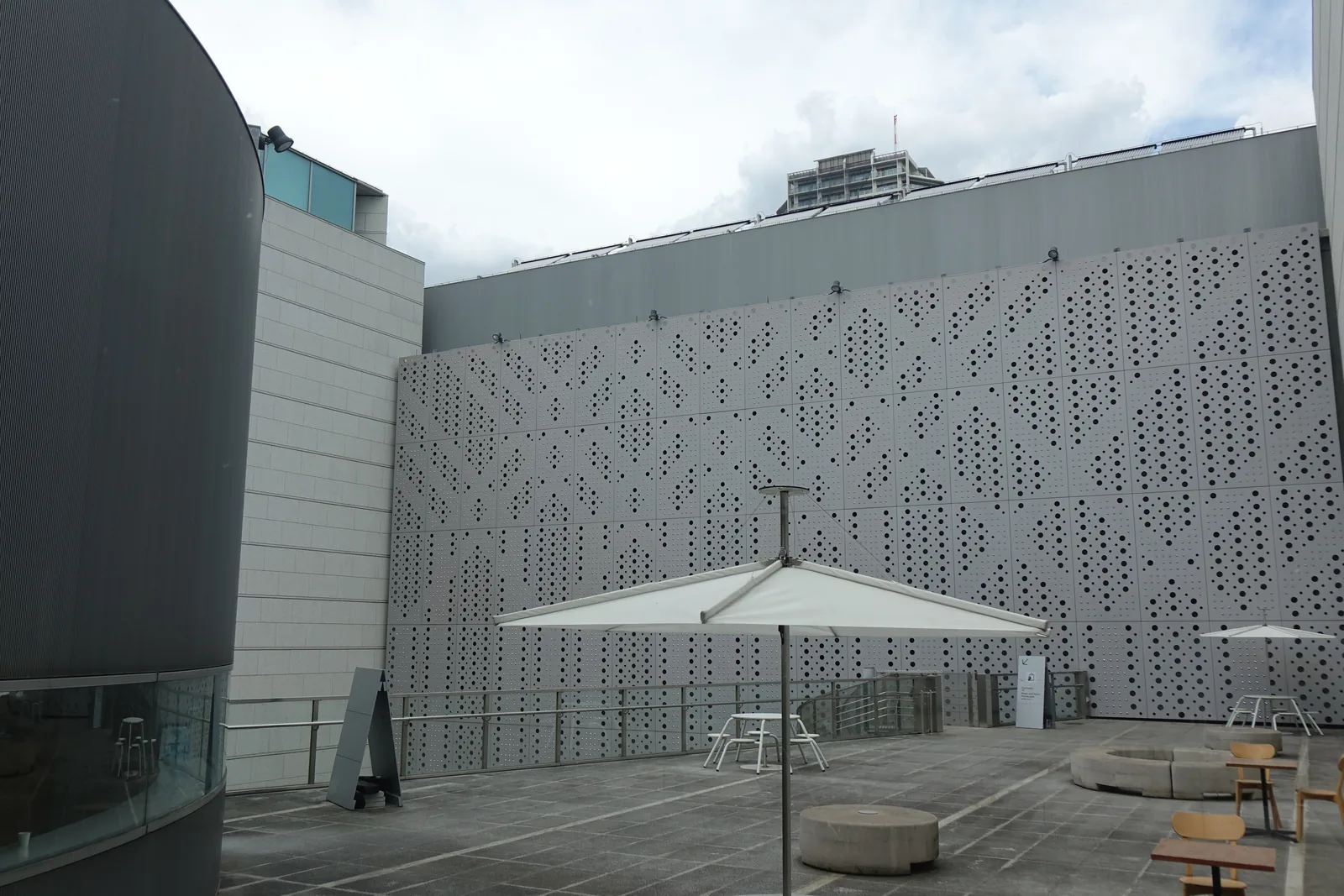 建築量體之間的中庭，一側為大片沖孔圖樣的淺灰色牆，左側為圓弧形深色量體，地坪上擺有白色遮陽傘、桌椅與圓形石凳