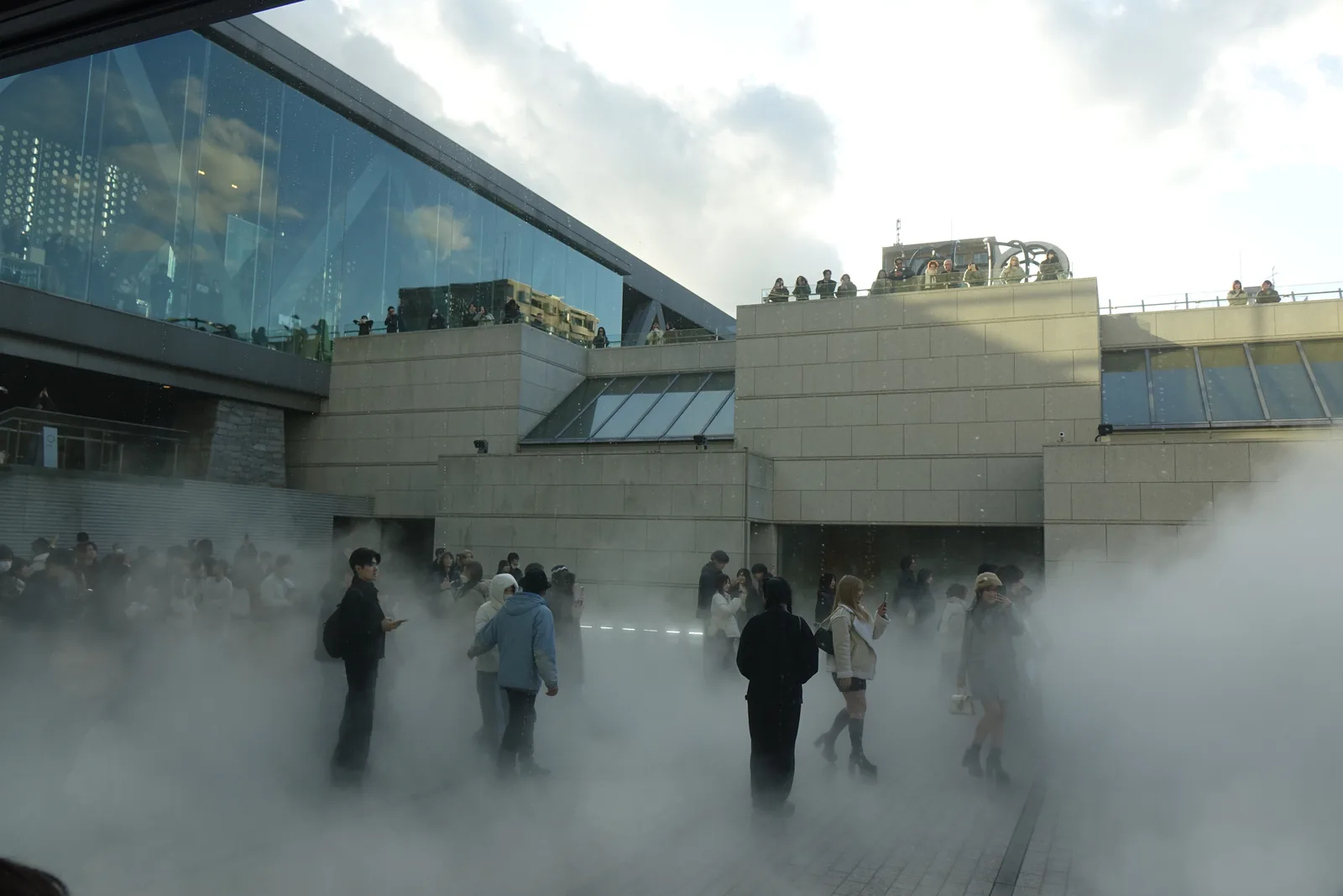 下沉庭園中瀰漫的人工霧，參觀者站在霧中，背景為層層退縮的石材量體與上方玻璃大廳的斜向屋頂