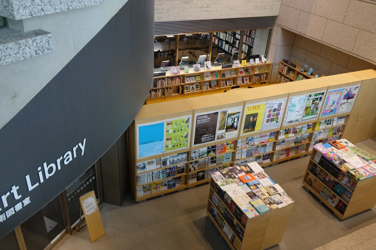 地下一樓美術圖書室入口，弧形深色牆面上標示Art Library，入口前為淺色木作的書報陳列架，擺放展覽摺頁與美術雜誌