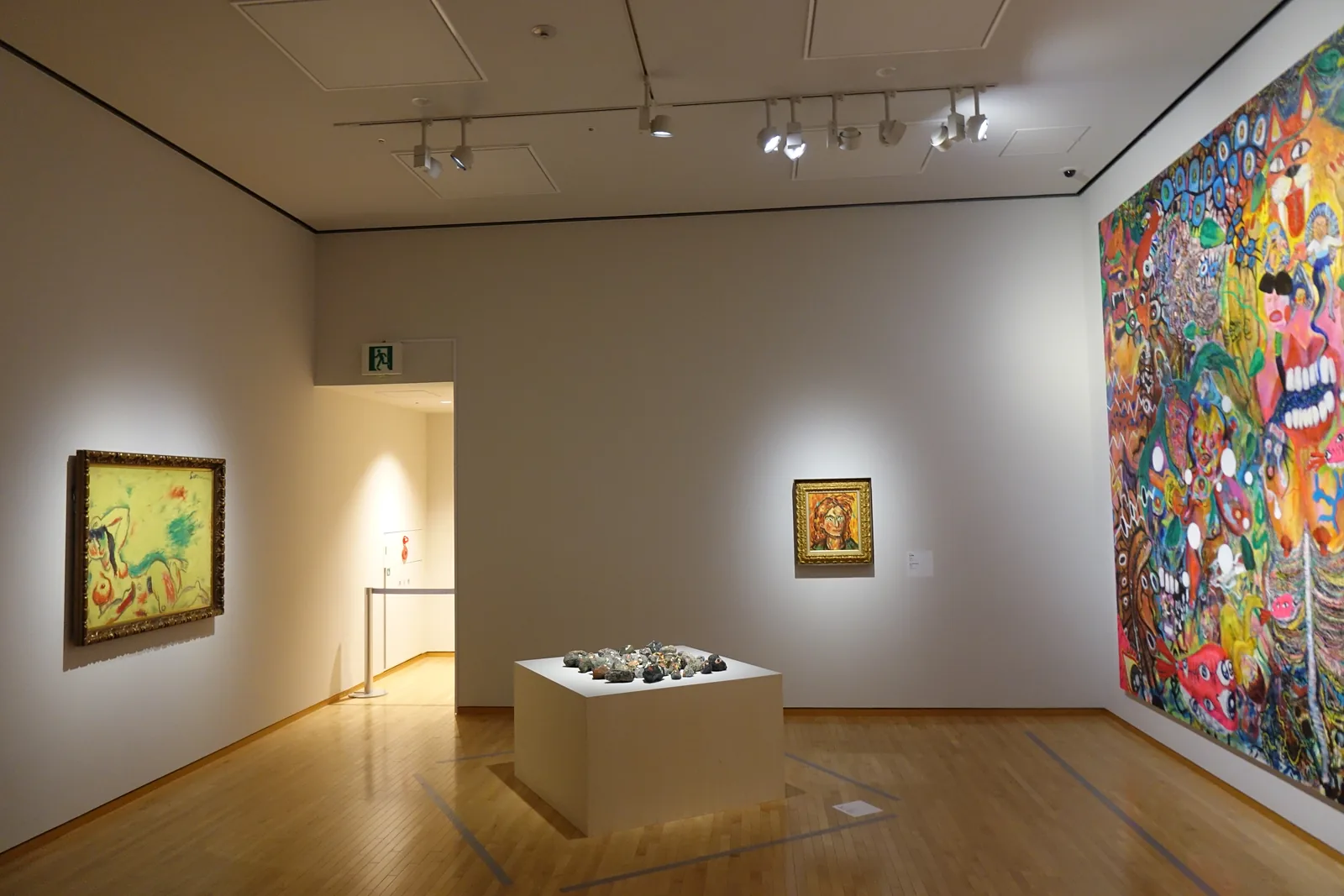 白色展示室，木地板上置有展示台，牆上掛著畫作，右側牆面為大幅色彩鮮豔的作品
