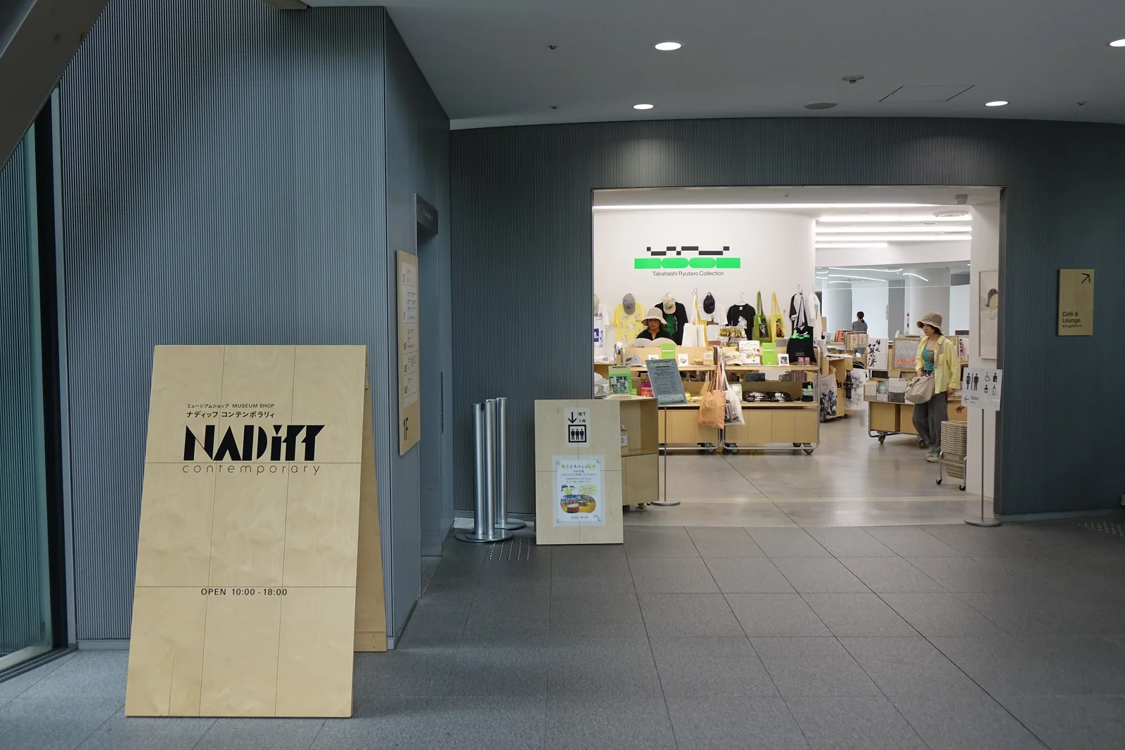 美術館商店入口，灰色牆面之間開出一處開口，前方立著木製看板，店內陳列商品