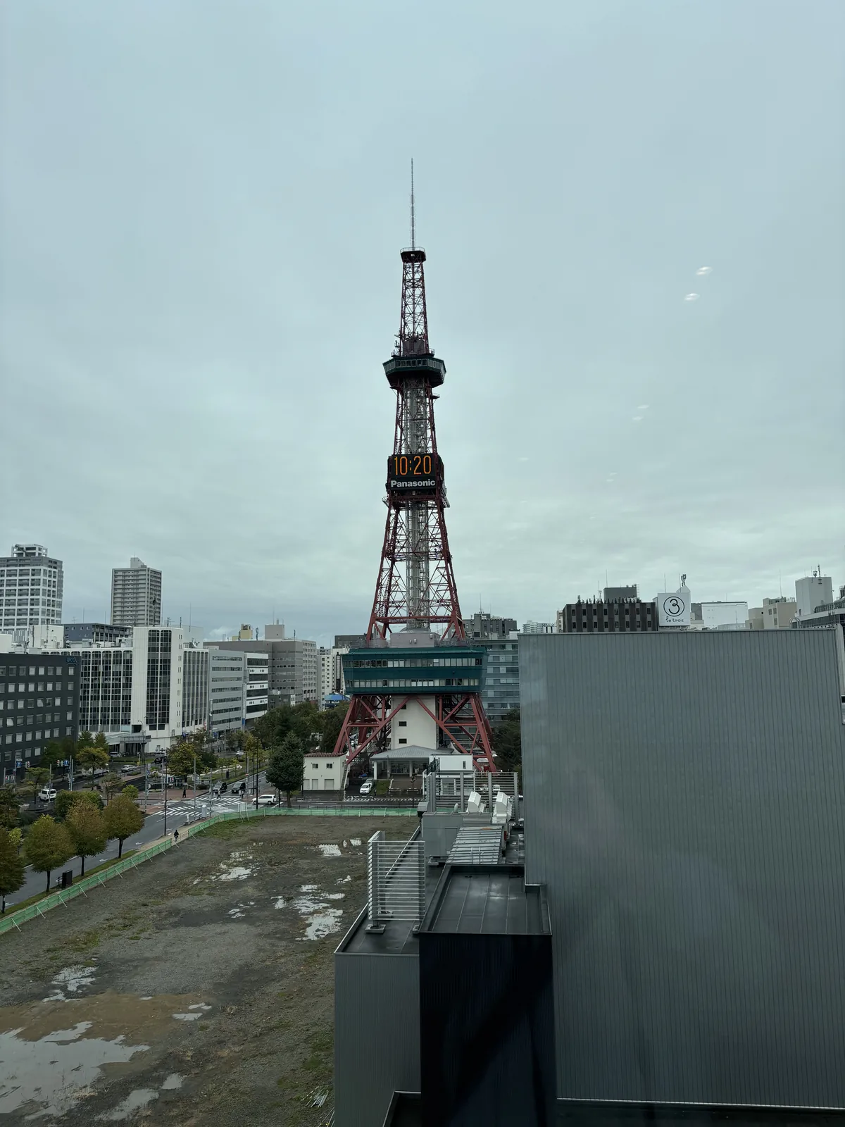 自四樓窗前望向札幌電視塔，前景為低層棟屋頂的深灰色設備量體，中景為一片空地與行道樹，電視塔鐵塔上的時鐘清晰可見