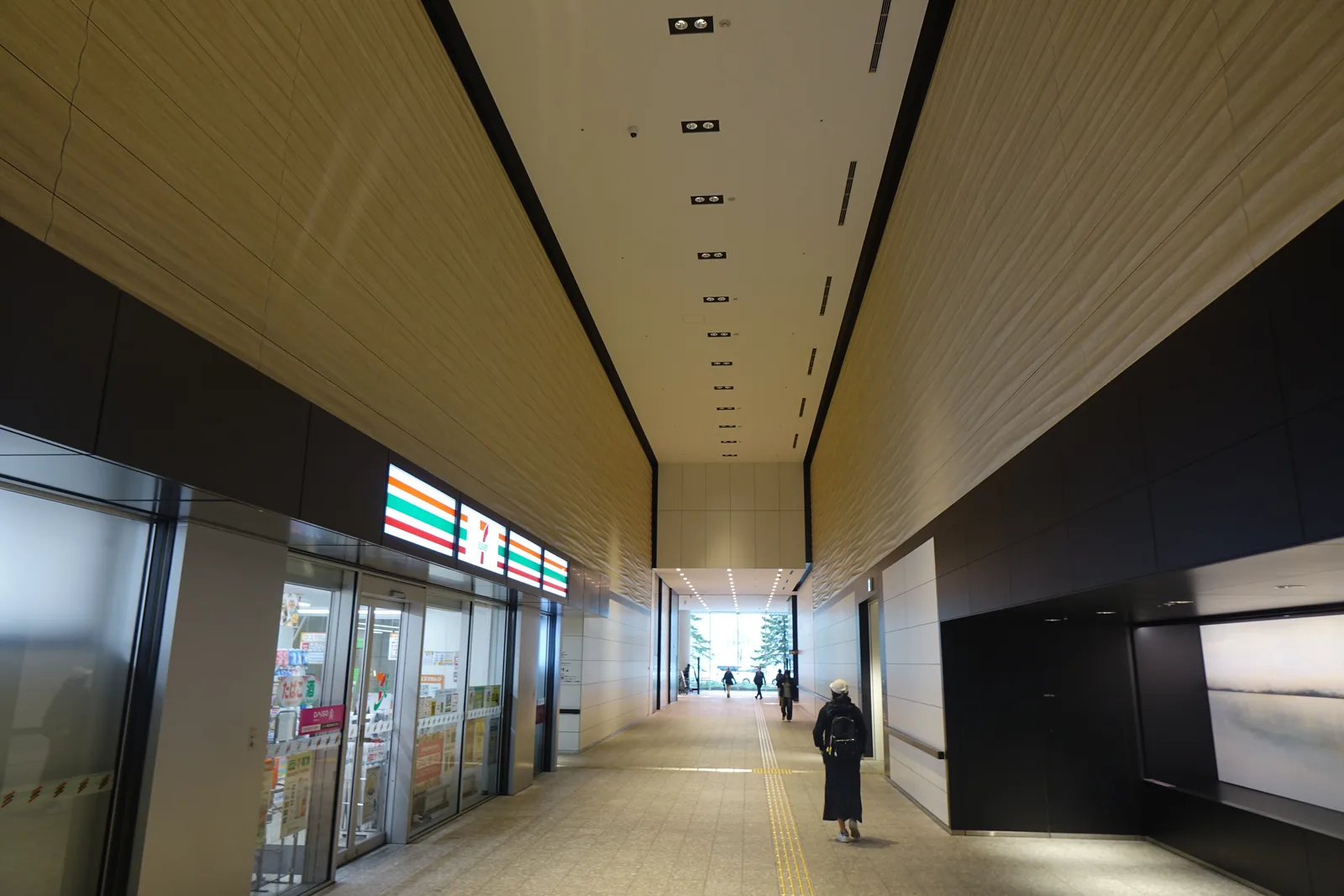 一樓貫穿建築的通道，左側為便利商店的玻璃門面，天花為淺色條板與一列筒燈，遠端為明亮的出口