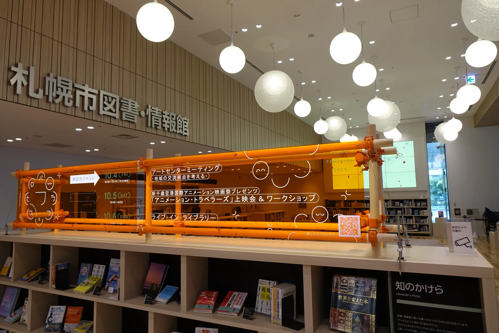 圖書・情報館一樓的書架，上方以橘色管材組成的框架懸掛活動資訊，後方牆面上標示館名，天花垂吊球形燈