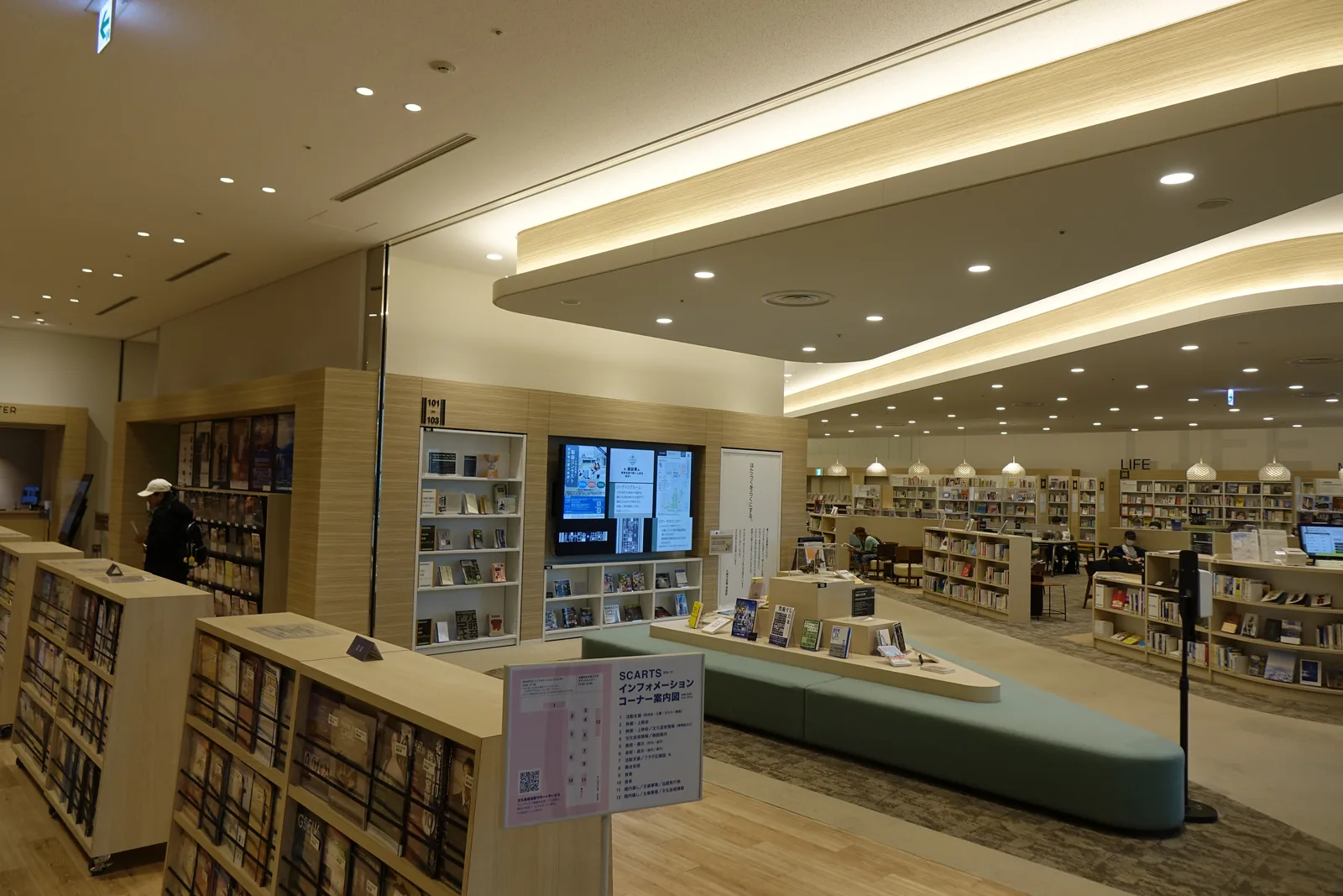 圖書・情報館二樓入口一帶，淺色木作書架與展示螢幕，曲線形的間接照明天花下為沙發座與閱覽區