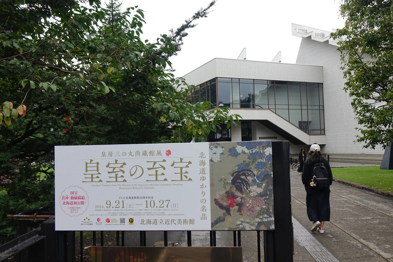 美術館入口前的展覽看板，後方為白色磁磚量體與懸挑的玻璃量體，右側為樹木，參觀者沿步道走向入口